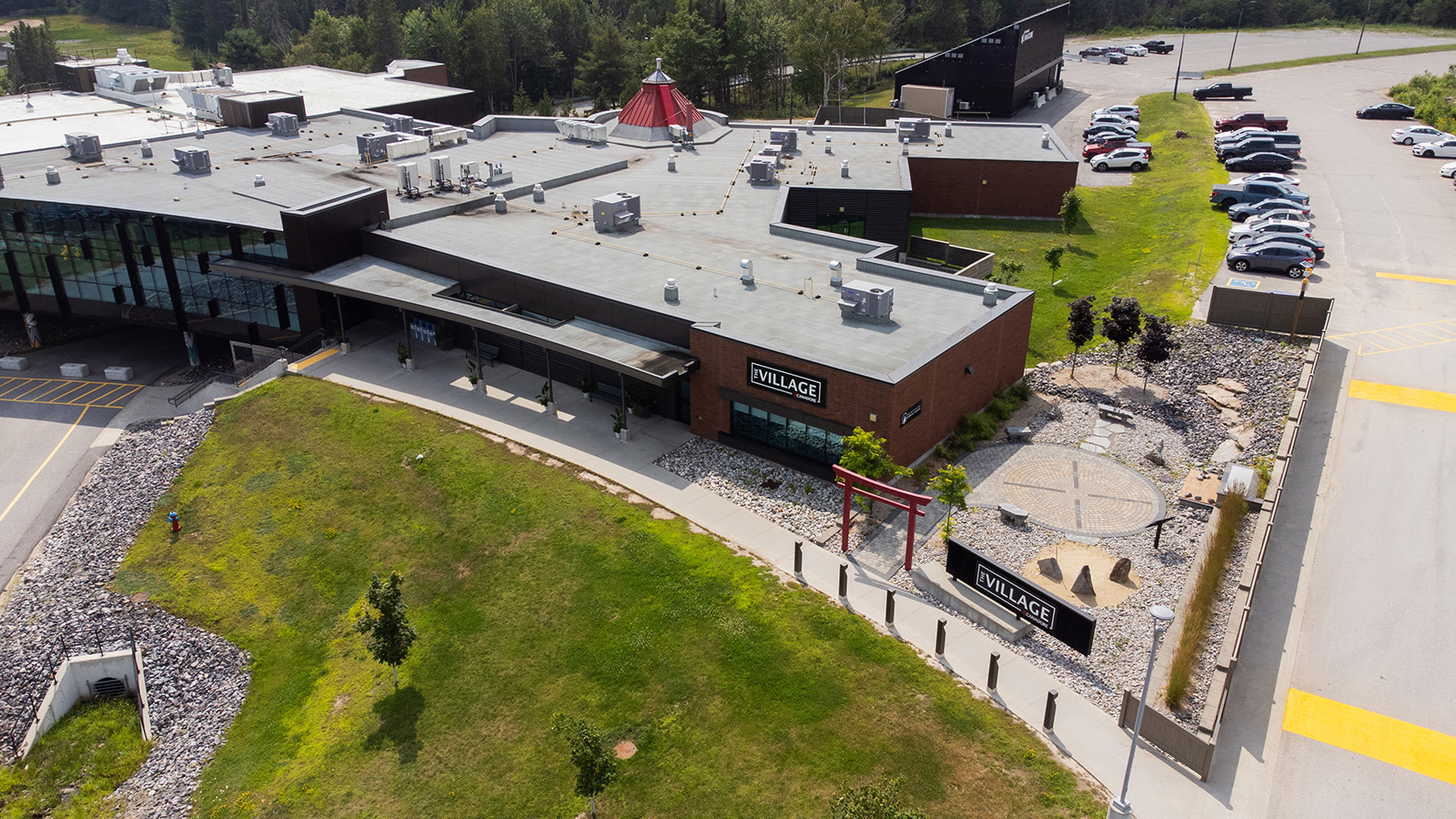 Aerial view of The Village, showcasing modern buildings with flat roofs, green spaces, parking lots, and a landscaped area featuring a red torii gate and rock garden.